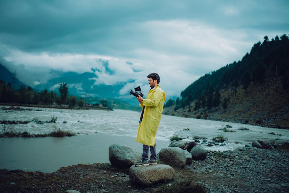 A man in a yellow raincoat stands by a river holding a camera, surrounded by scenic nature.