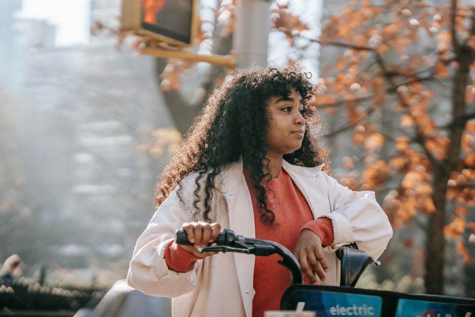 Dreamy young black lady in casual outfit looking away on street with bike in town in sunny day near tree with orange leaves and traffic light