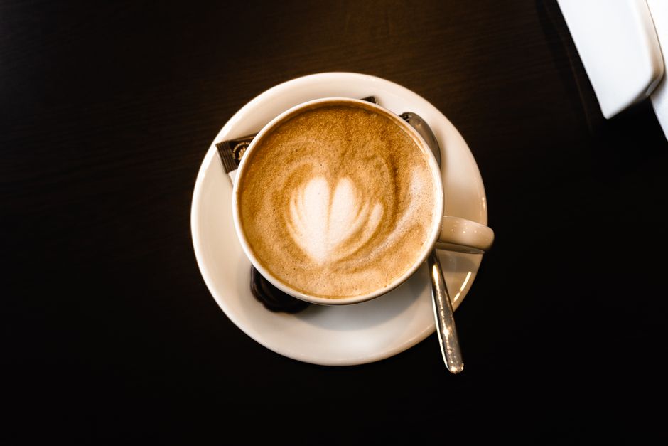 Overhead shot of a latte in a white cup with heart-shaped foam art on a dark table.
