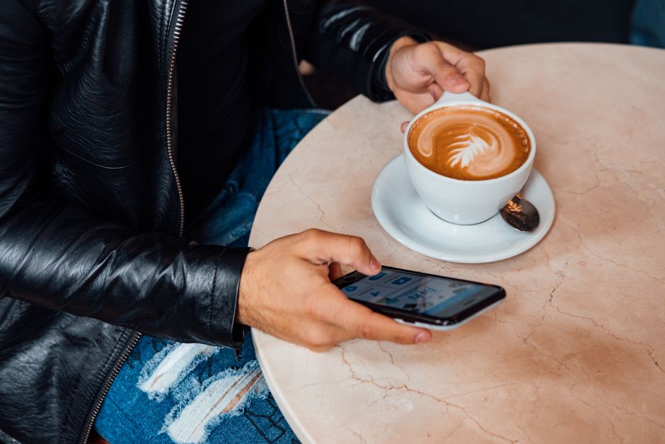 Person enjoying a latte art coffee while using a smartphone, wearing a black leather jacket.