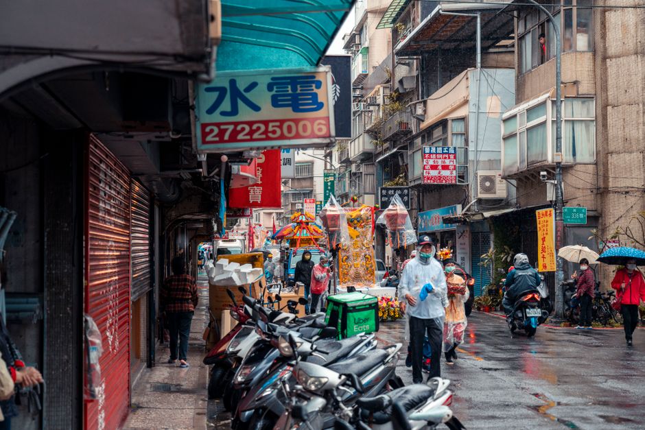 Lively Asian city street scene with people, scooters, and colorful signs on a rainy day.