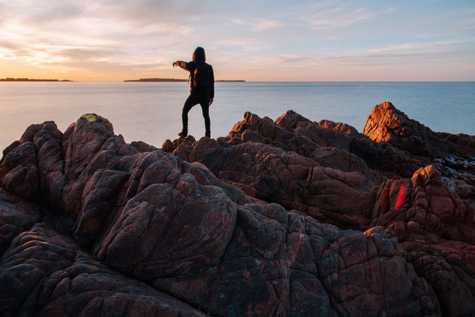 Back view of unrecognizable traveller in warm wear standing on rocky seashore and pointing at beautiful sky