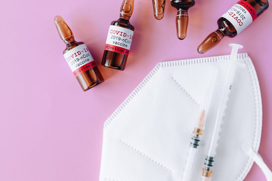 Close-up of COVID-19 vaccine vials, syringe, and mask on a pink surface.